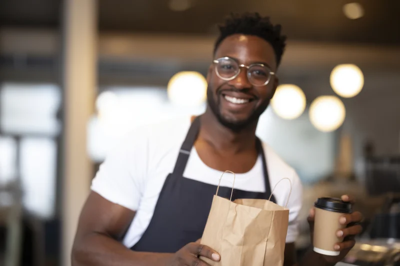 Smiling black barista holding paper bag and takeout coffee cup in cafe