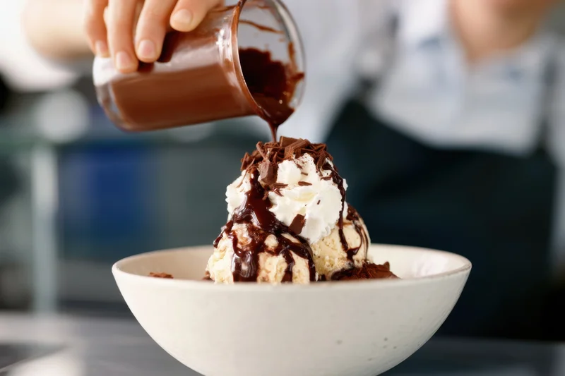 Barista pouring chocolate sauce over ice cream with whipped cream and chocolate shavings in a bowl