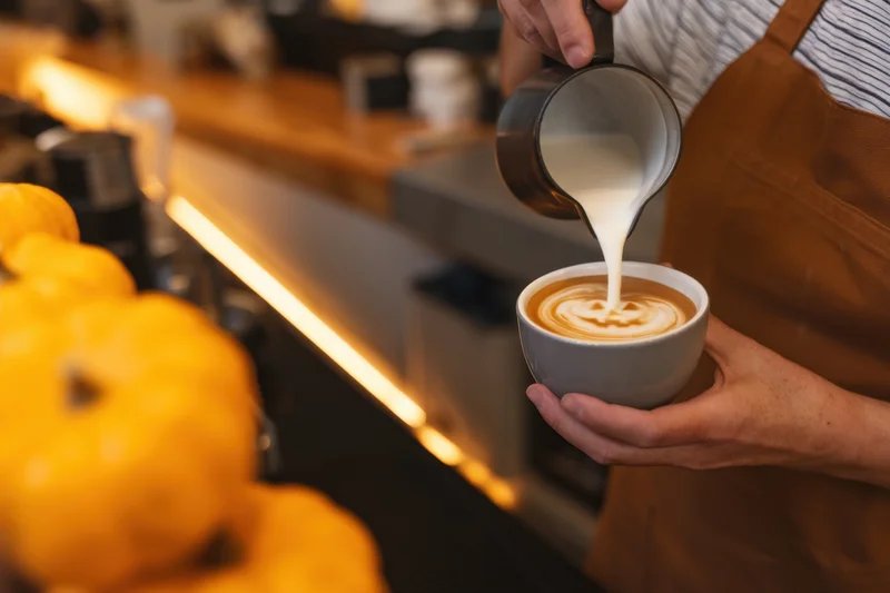 Barista pouring steamed milk into a latte with pumpkin-themed latte art in a cafe setting