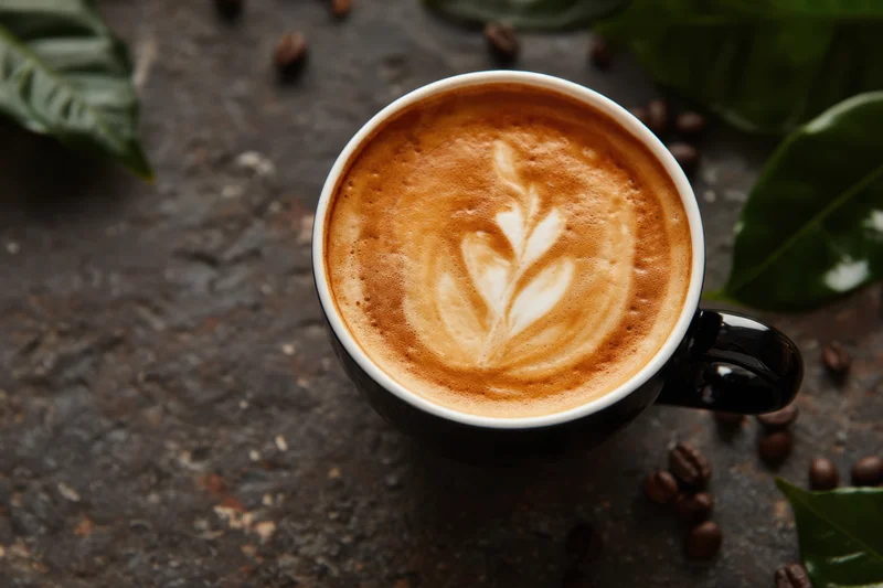 Latte art in a black ceramic cup with coffee beans and green leaves on a dark stone surface