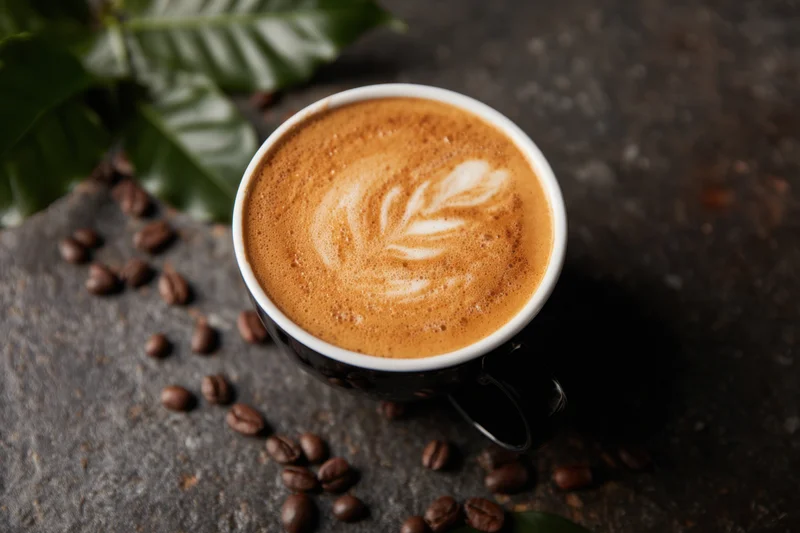 Latte art in a black ceramic cup with coffee beans and green leaves on a dark surface