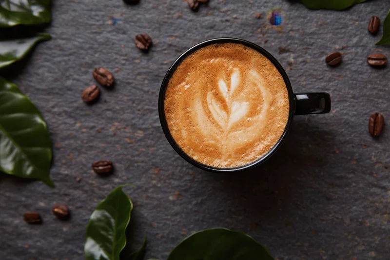 Latte art in a black ceramic cup with coffee beans and green leaves on a dark stone surface, top view