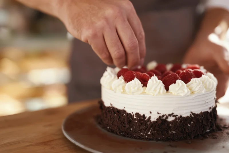 Baker garnishing a raspberry cream cake with chocolate flakes on a wooden board