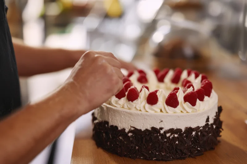 Baker garnishing a raspberry cream cake with chocolate flakes on a wooden table