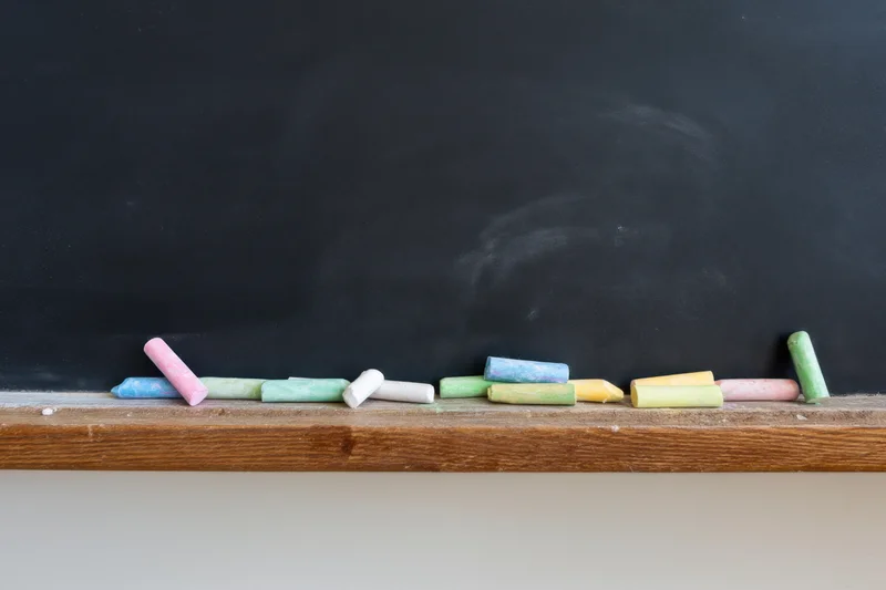 Colorful chalk pieces scattered on wooden ledge of black chalkboard in classroom