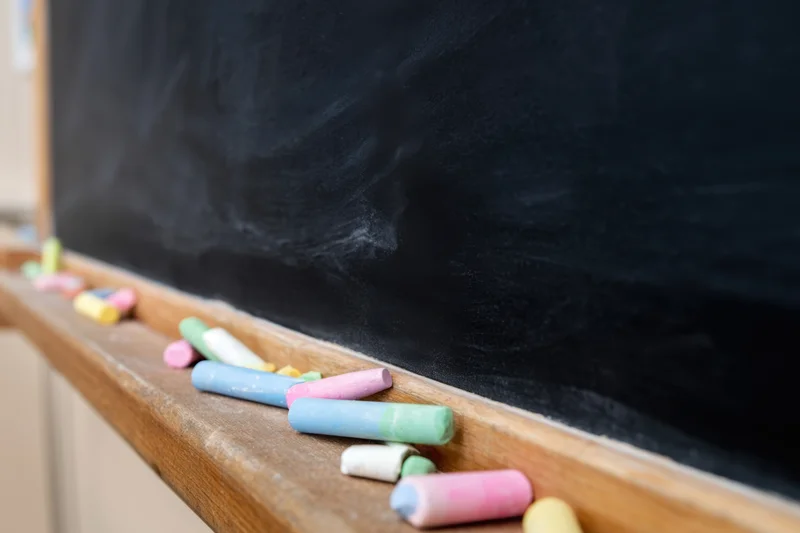Colorful chalk pieces scattered on wooden ledge of black chalkboard in classroom