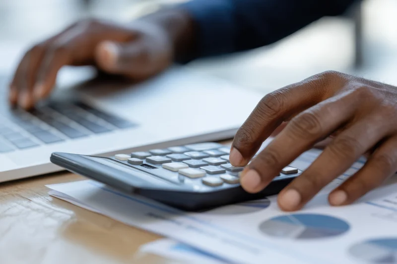 Hands calculating on a calculator and typing on a laptop at a desk with financial documents