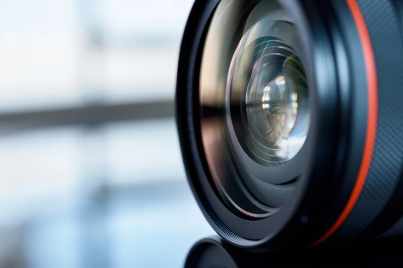 Closeup of a camera lens showing glass elements and red rim against a blurred background