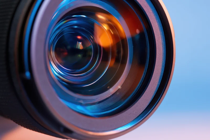Closeup of a camera lens showing intricate glass elements and colorful reflections against a gradient background