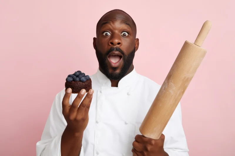 Baker in white coat holding blueberry cake and rolling pin against pink background