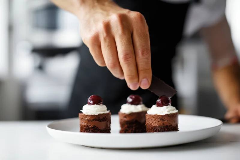 Chef decorating three chocolate desserts with whipped cream and cherries on a white plate