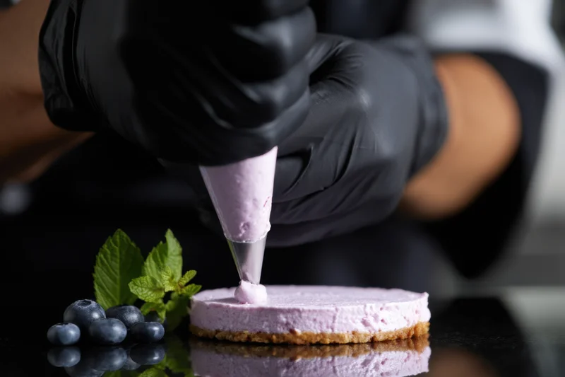 Chef's hands in black gloves piping lavender cream onto a small cake with blueberries and mint nearby on a reflective surface