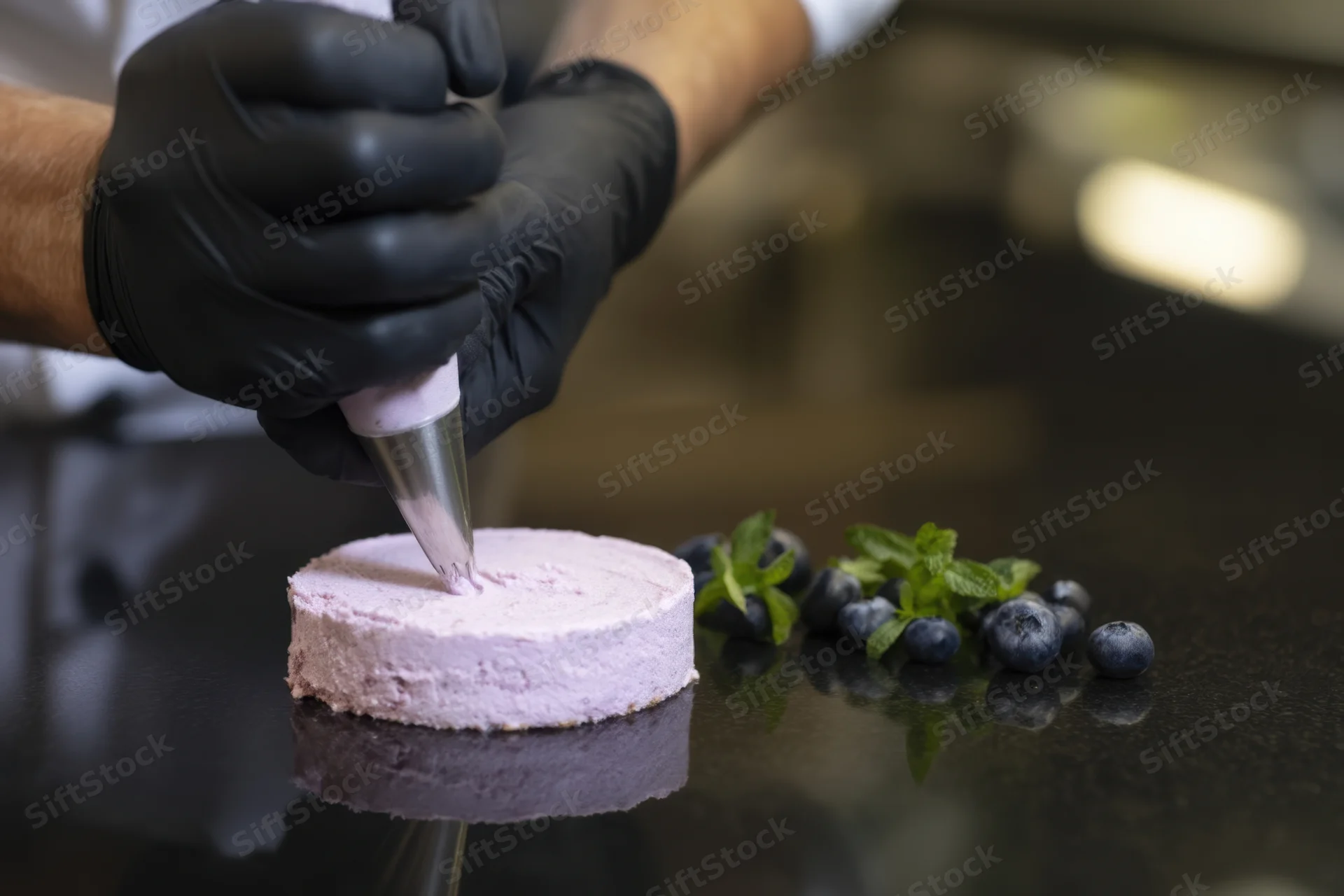 Chef in black gloves piping lavender frosting onto a small cake with blueberries and mint nearby on a dark countertop