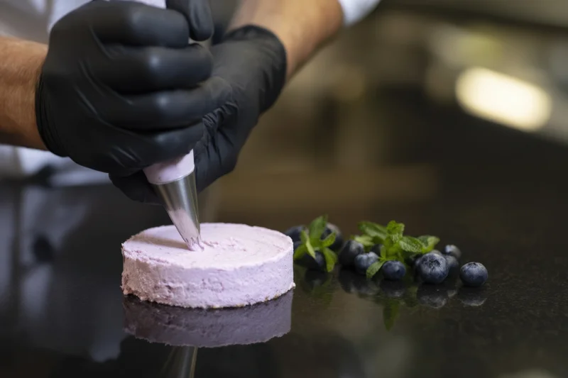 Chef in black gloves piping lavender frosting onto a small cake with blueberries and mint nearby on a dark countertop
