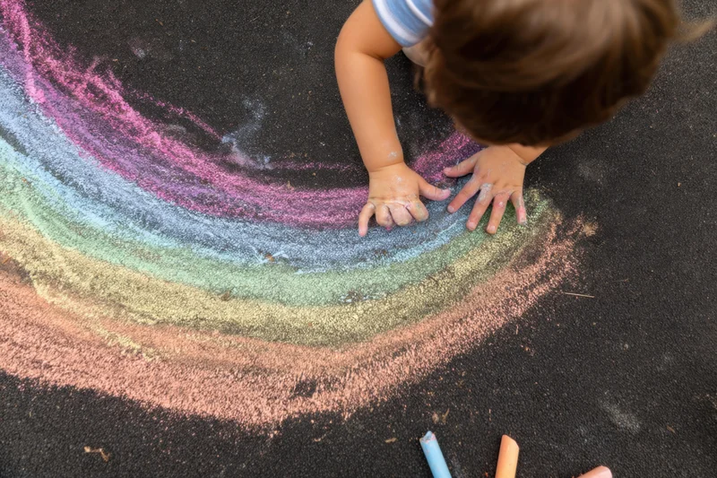 Child drawing a rainbow with colorful chalk on black pavement