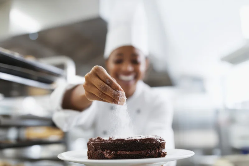 Female pastry chef sprinkling powdered sugar on chocolate cake in bright kitchen