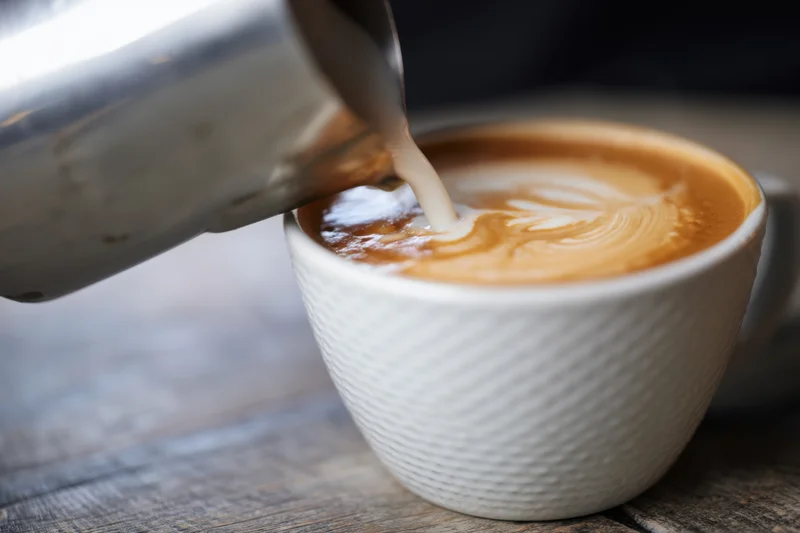 Milk being poured into a latte to create latte art in a white ceramic cup on a wooden table