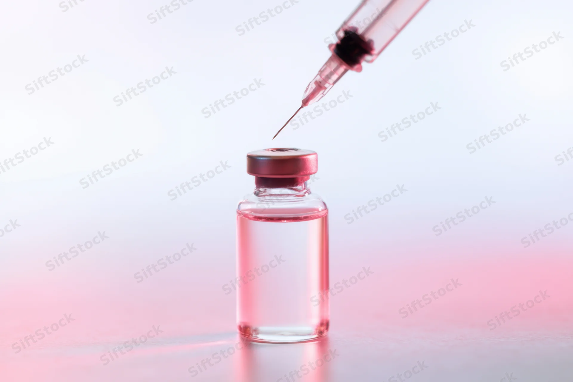 Glass vial with pink-tinted liquid and syringe needle poised above it on a smooth surface
