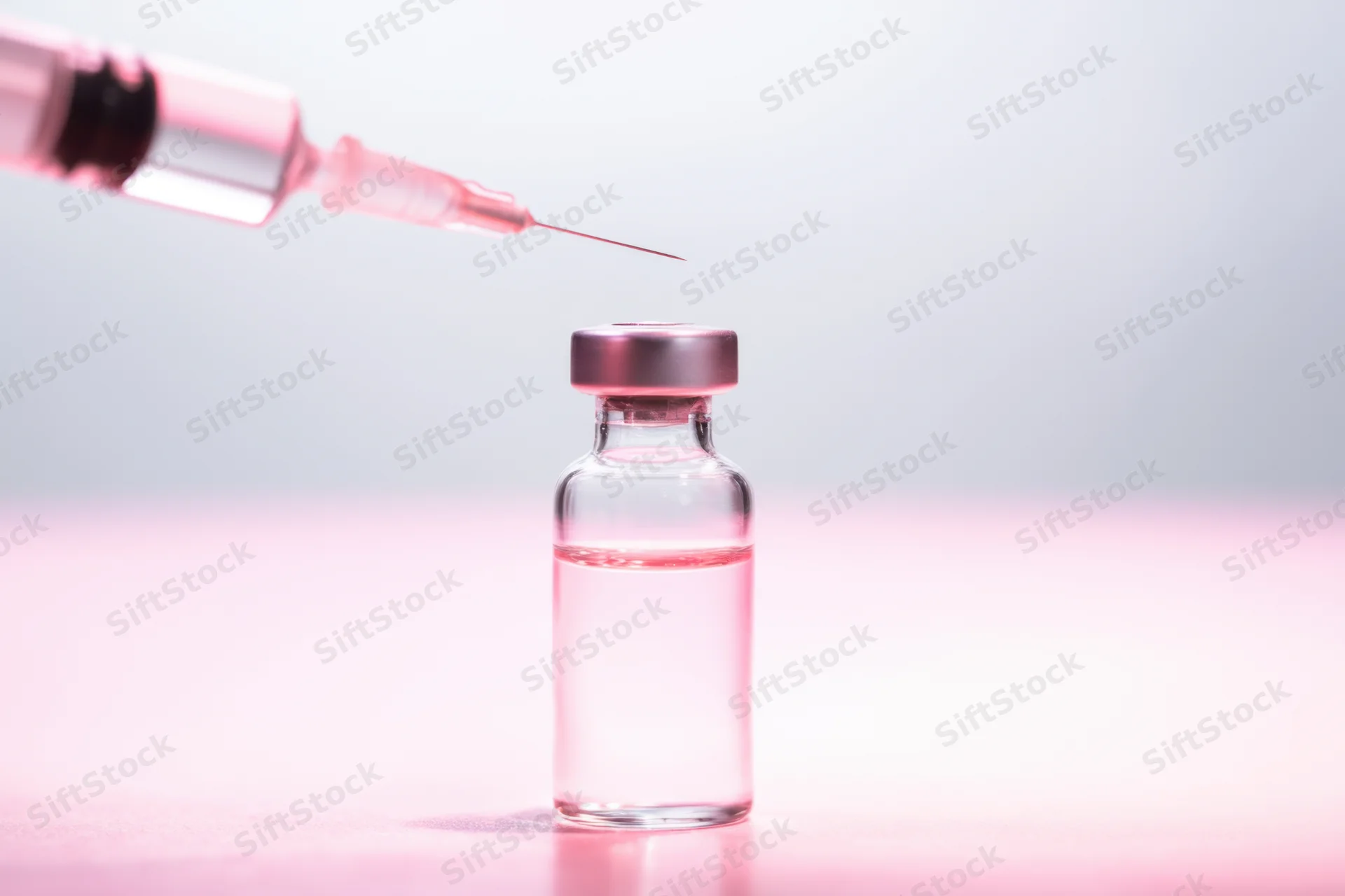 Glass vial with pink-tinted liquid and syringe poised above it on a pink surface