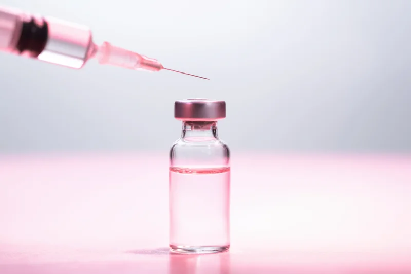 Glass vial with pink-tinted liquid and syringe poised above it on a pink surface