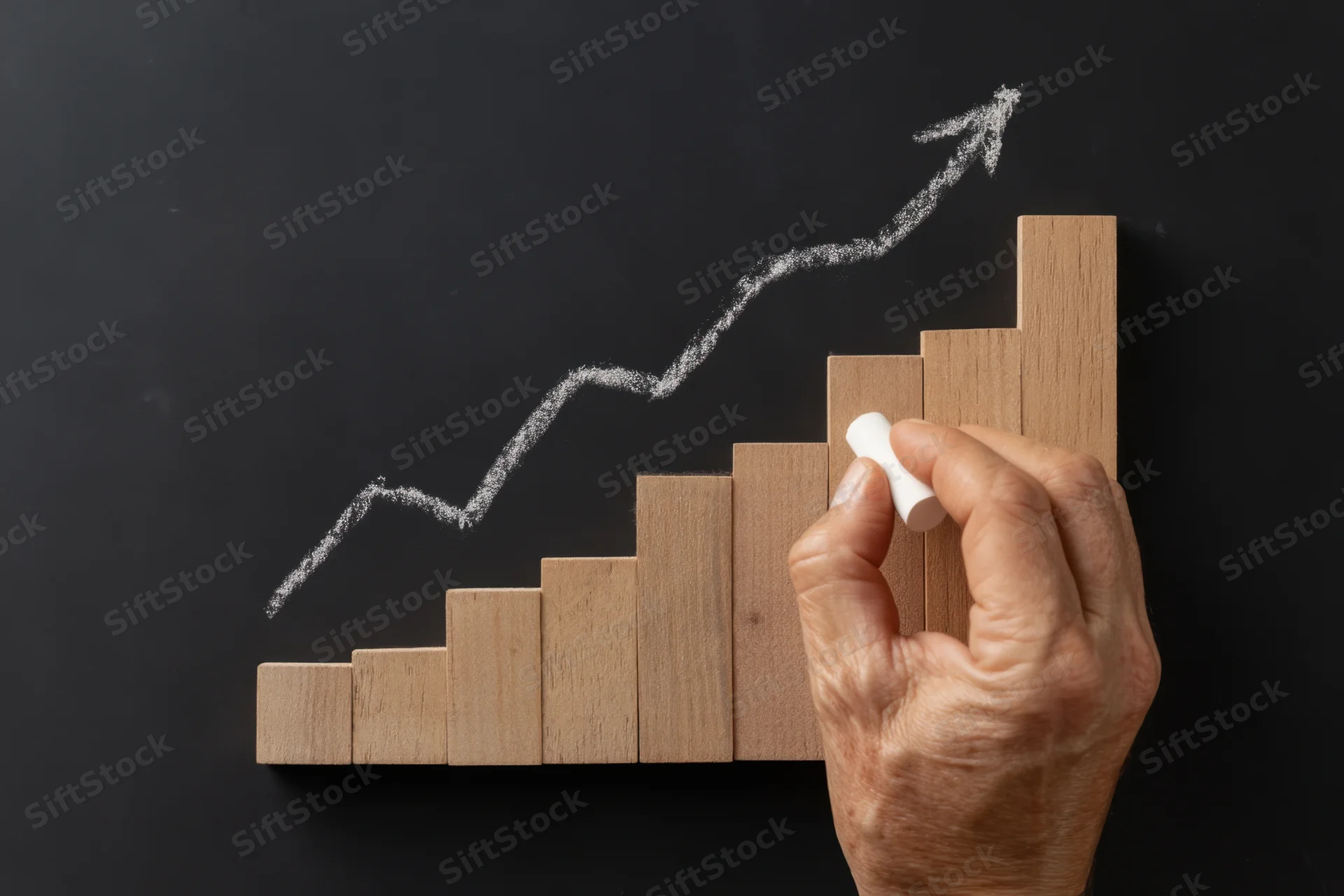 Hand drawing upward arrow on blackboard beside wooden blocks arranged as growth chart