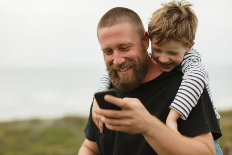 Father carrying son on shoulders while both smile at smartphone outdoors near ocean