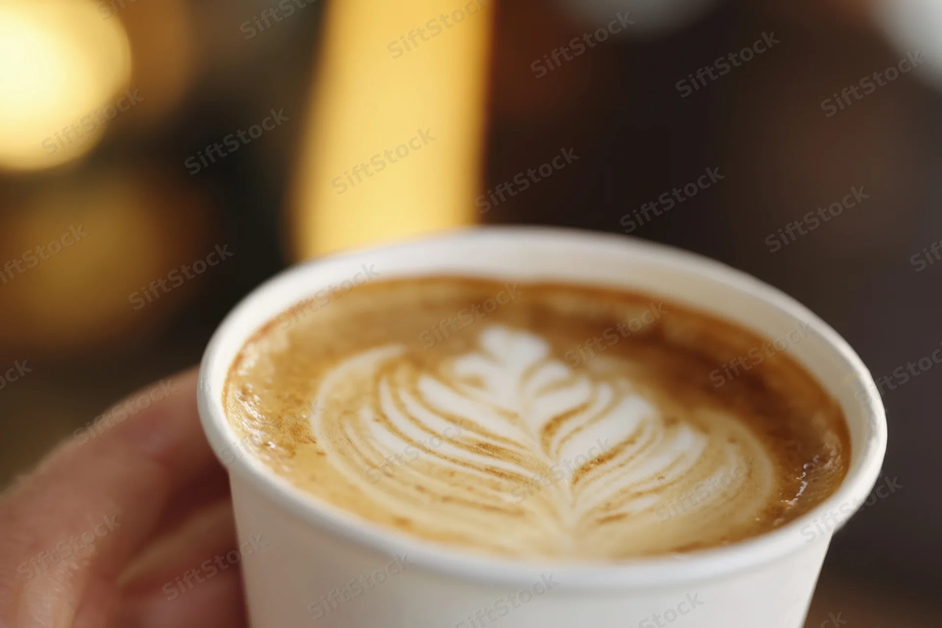 Hand holding a latte in a white disposable cup with leaf-shaped foam art, close-up view against blurred warm background
