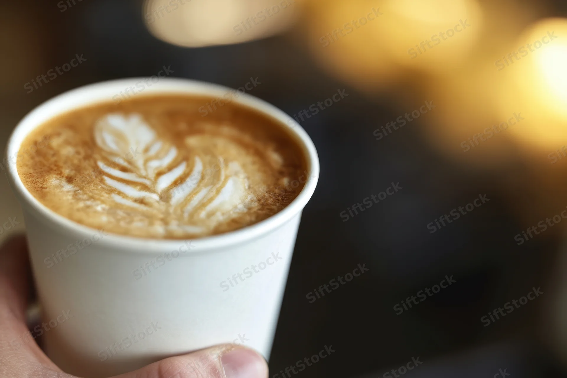 Hand holding a latte in a white disposable cup with leaf latte art, soft bokeh background