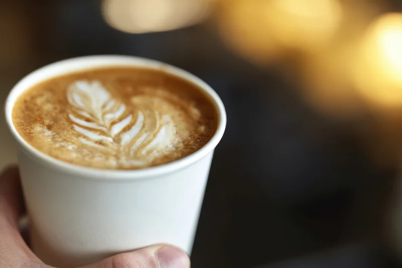 Hand holding a latte in a white disposable cup with leaf latte art, soft bokeh background