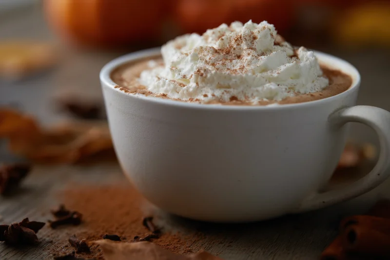 Latte topped with whipped cream and cinnamon dusted on a wooden table with autumn spices scattered around