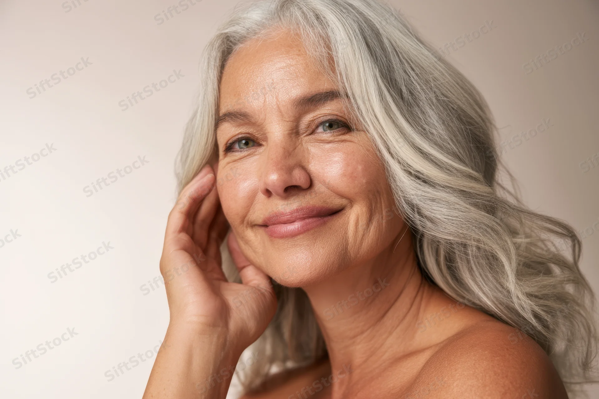 Mature woman with long wavy silver hair smiling gently with hand on cheek in studio portrait