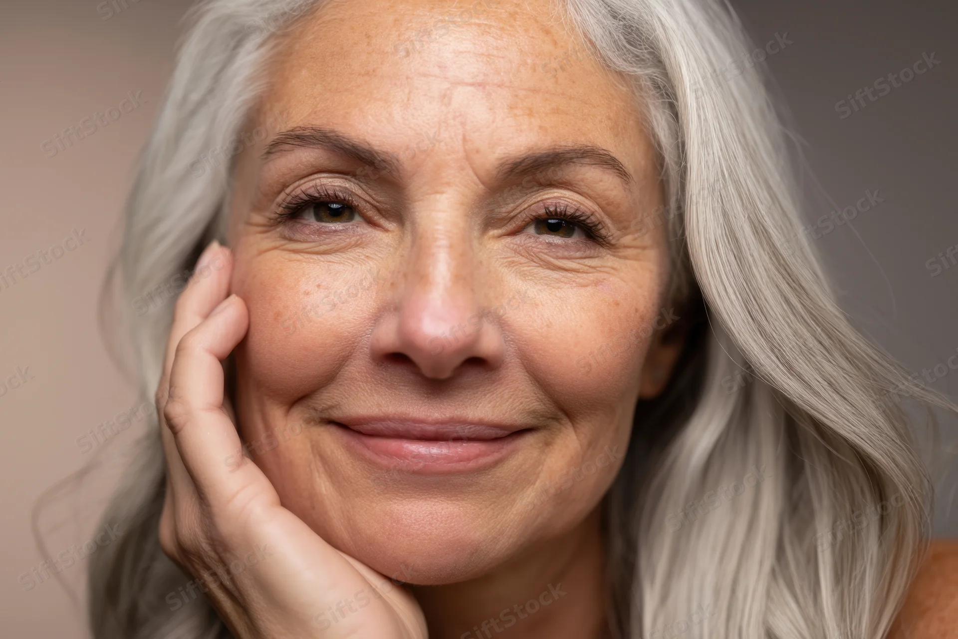 Mature woman with long wavy silver hair smiling gently with hand on cheek in studio portrait