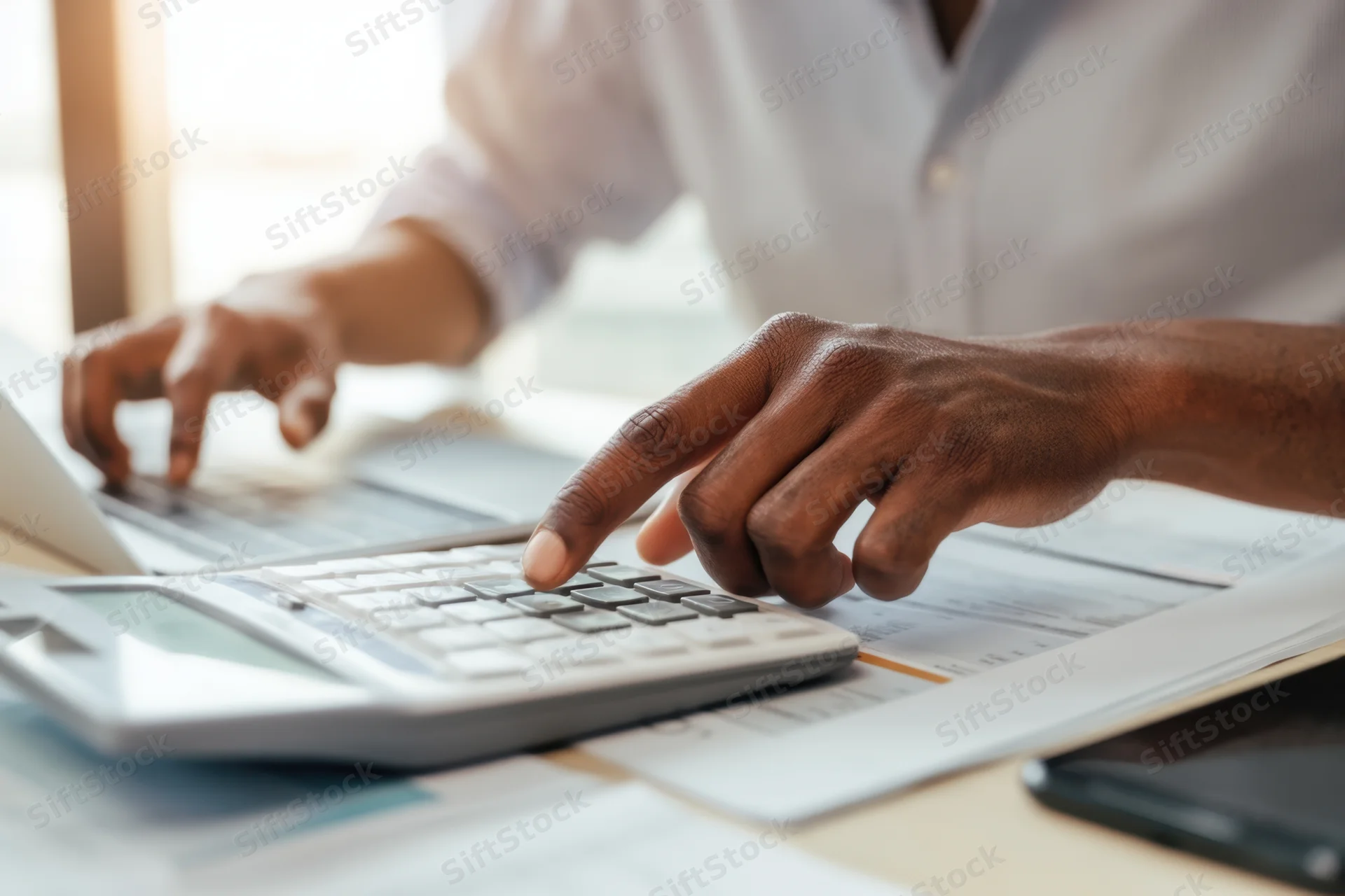 Black man calculating figures on a desk with calculator and laptop in modern office workspace