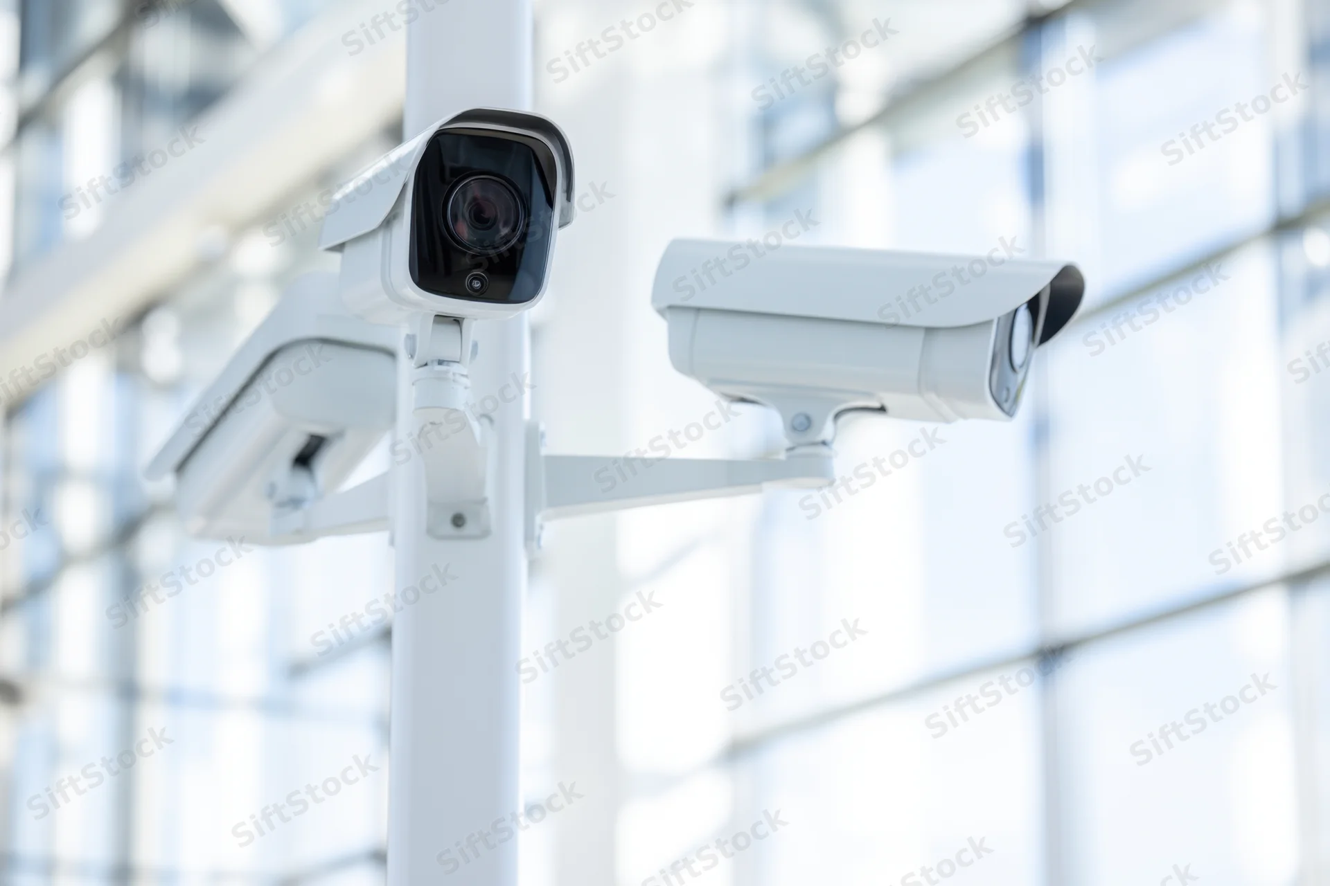Three white security cameras mounted on a pole outside a modern glass building