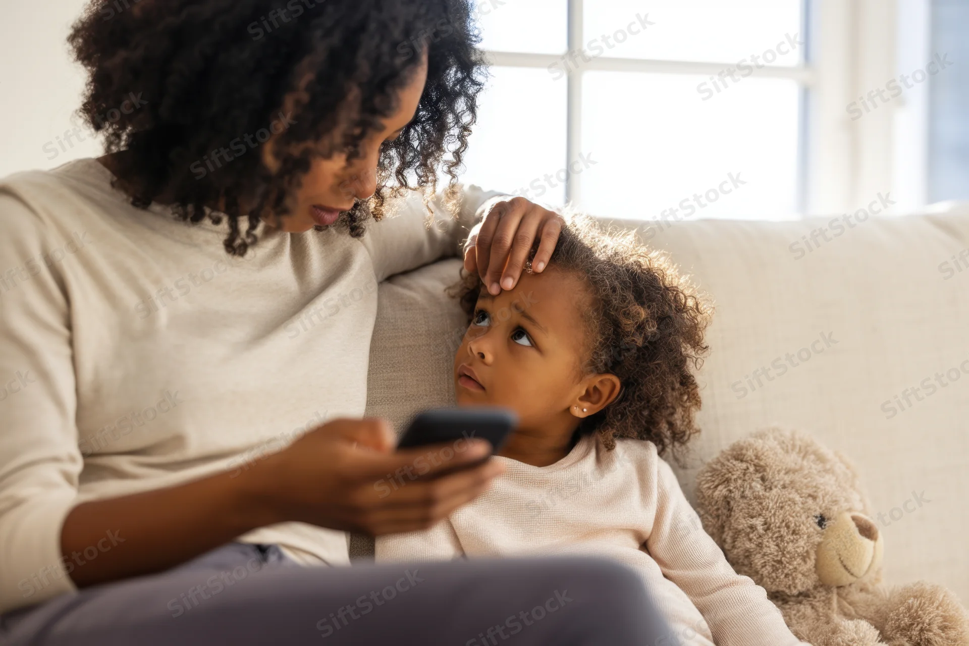 Mother checking smartphone while gently touching her daughter’s head on a couch at home