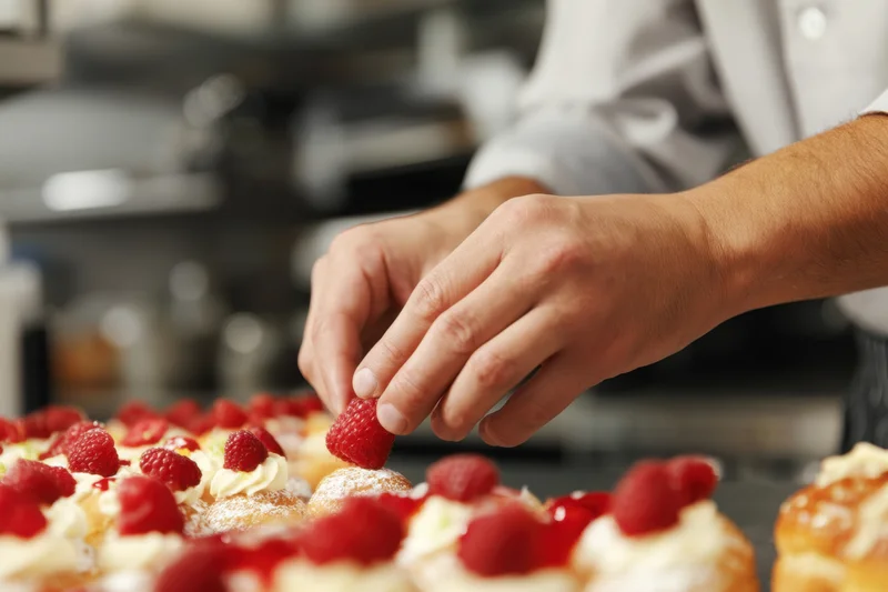Pastry chef placing fresh raspberries atop cream-topped pastries in a kitchen