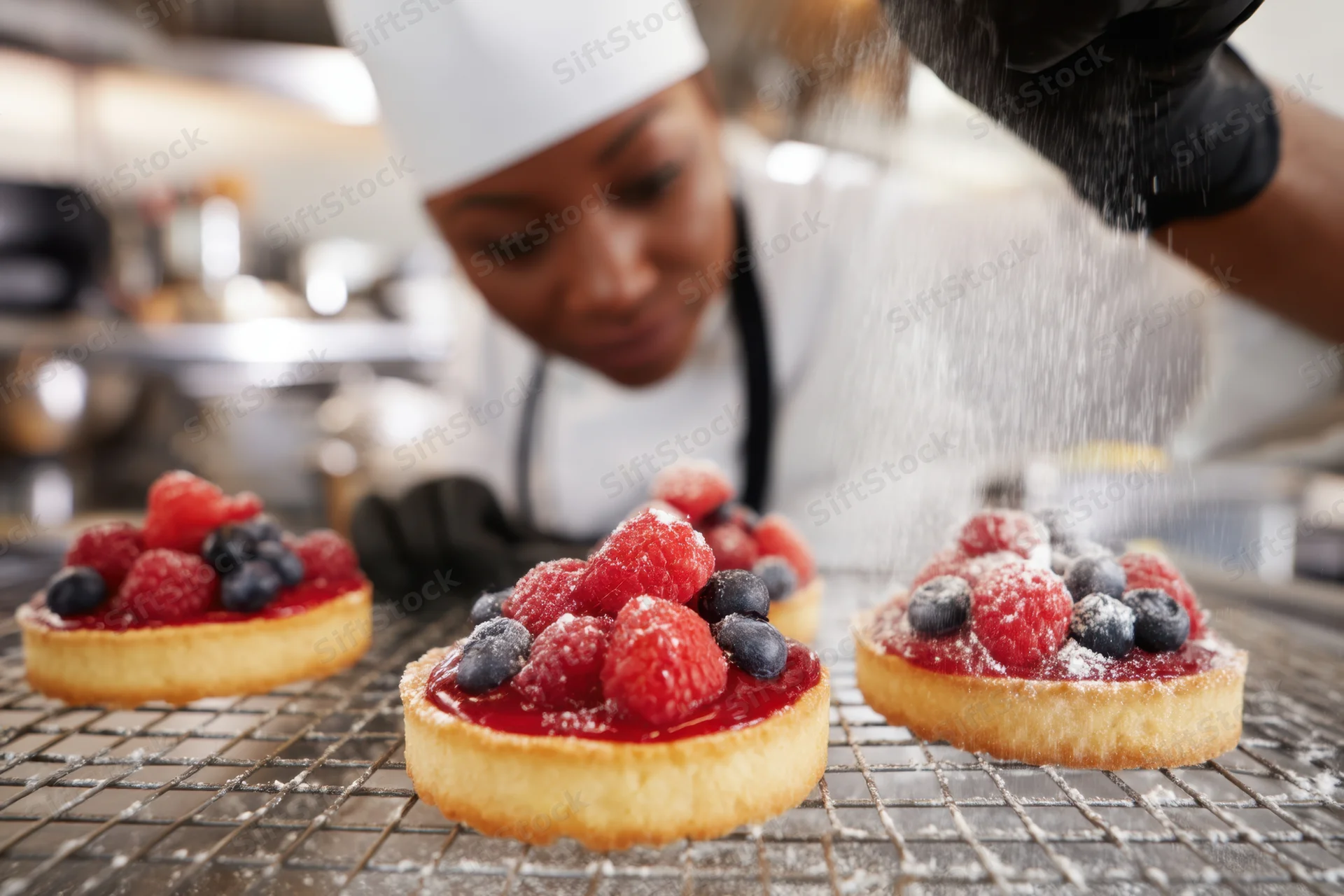 Pastry chef sprinkling powdered sugar on fruit tarts arranged on a cooling rack in a kitchen