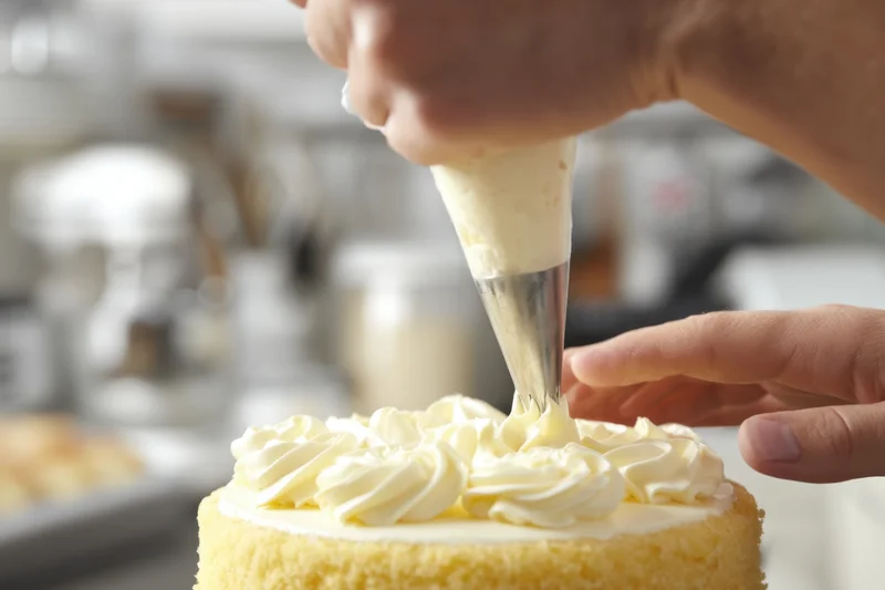 Pastry chef piping cream frosting onto a yellow cake in a kitchen