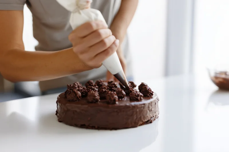 Woman decorating chocolate cake with piping bag on white countertop