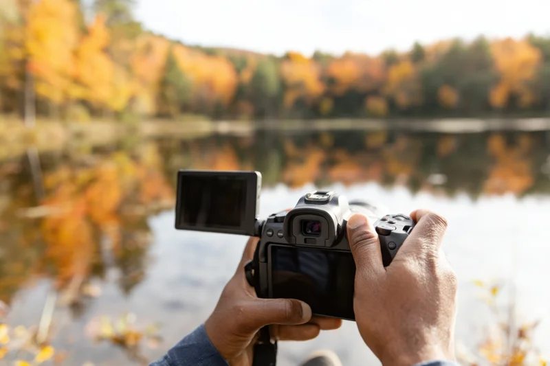 Photographer holding mirrorless camera with flip screen near autumn lake with colorful foliage reflection in water