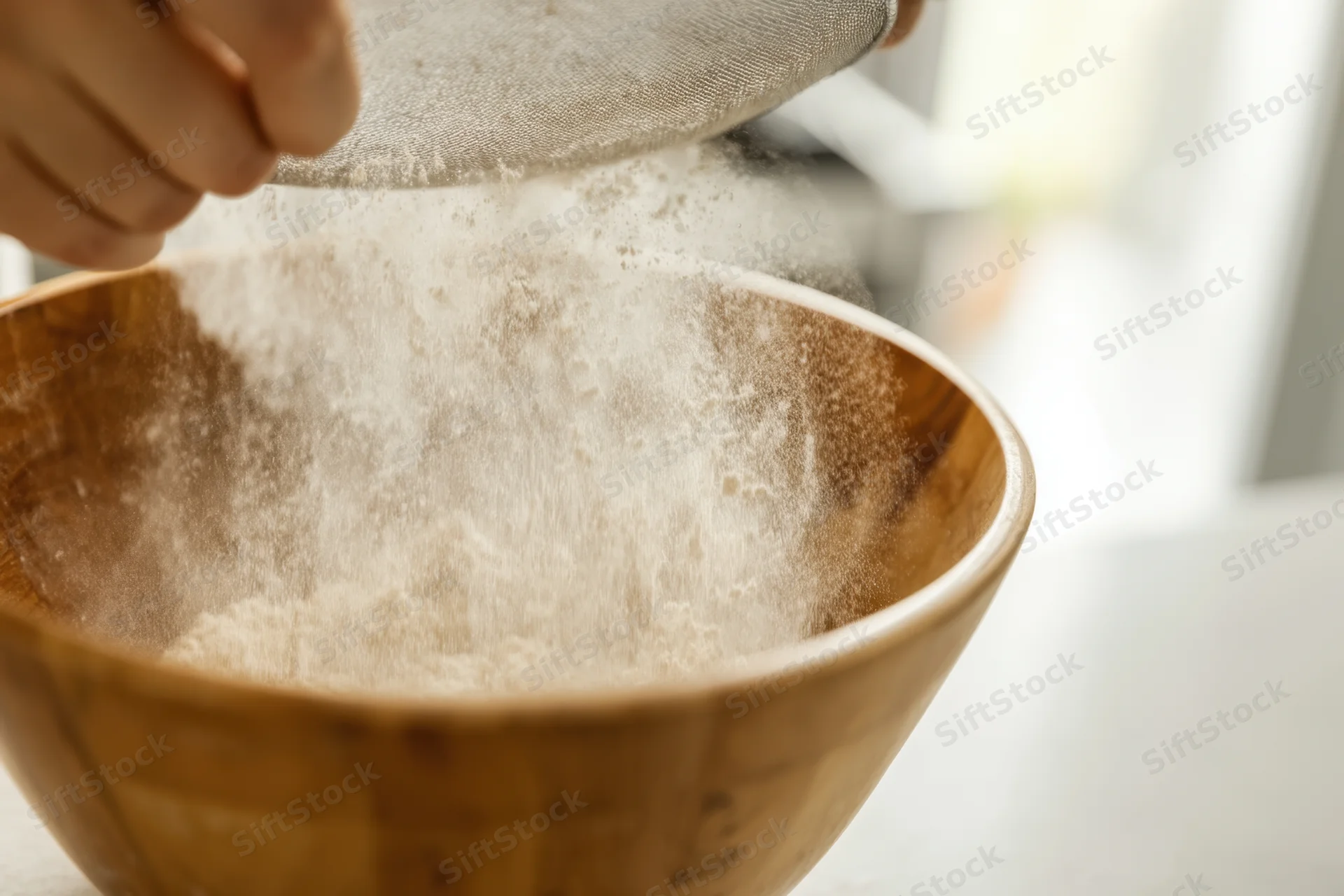 A person sifting flour through a fine-mesh sieve into a wooden bowl in a kitchen