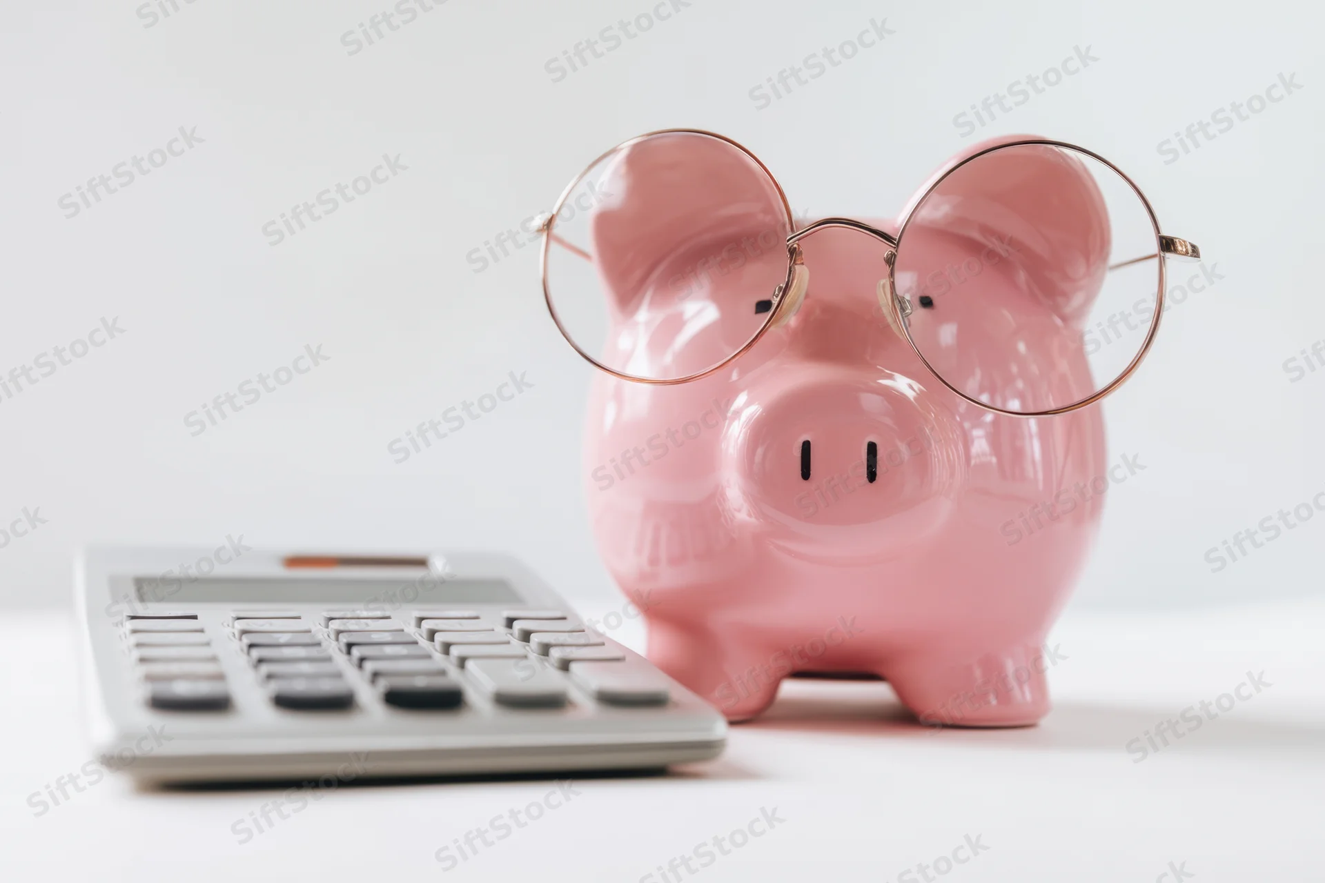 Pink piggy bank wearing round glasses beside a calculator on a white surface