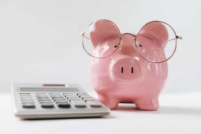 Pink piggy bank wearing round glasses beside a calculator on a white surface