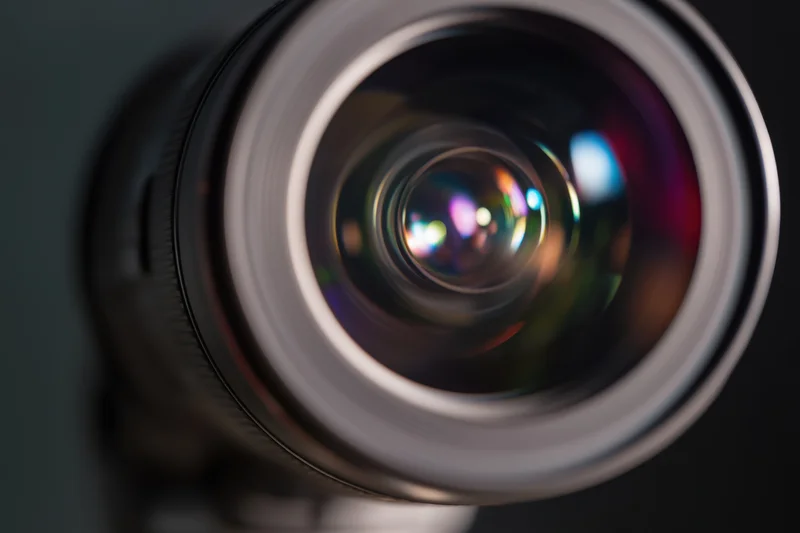 Closeup of a professional camera lens showing intricate glass elements with colorful reflections in a dark setting