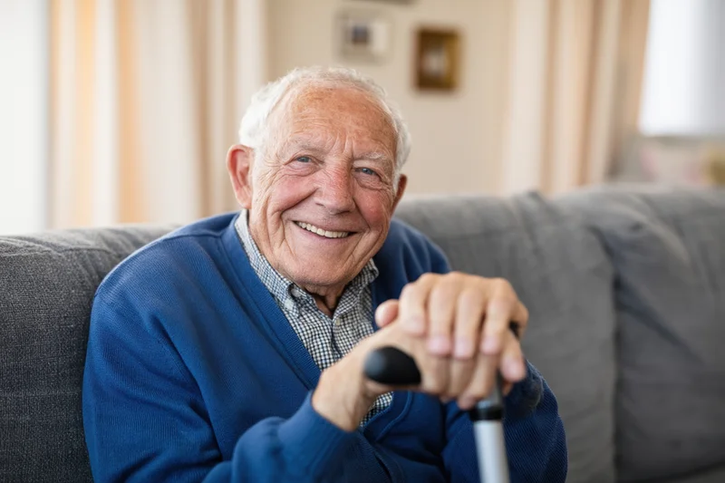 Senior man smiling while sitting on a gray sofa holding a cane in a living room