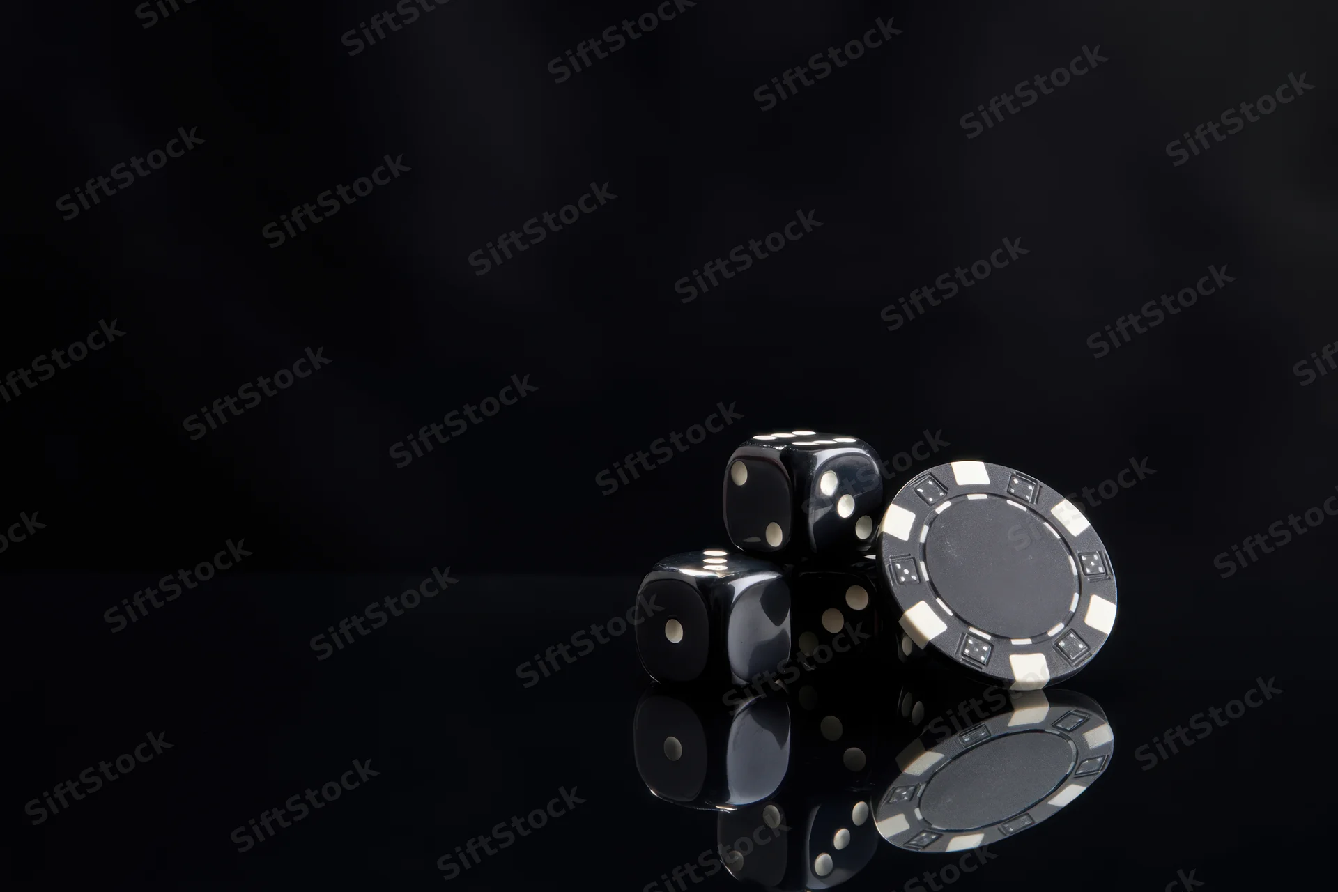 Black casino dice and a poker chip arranged on a glossy reflective surface with dark background
