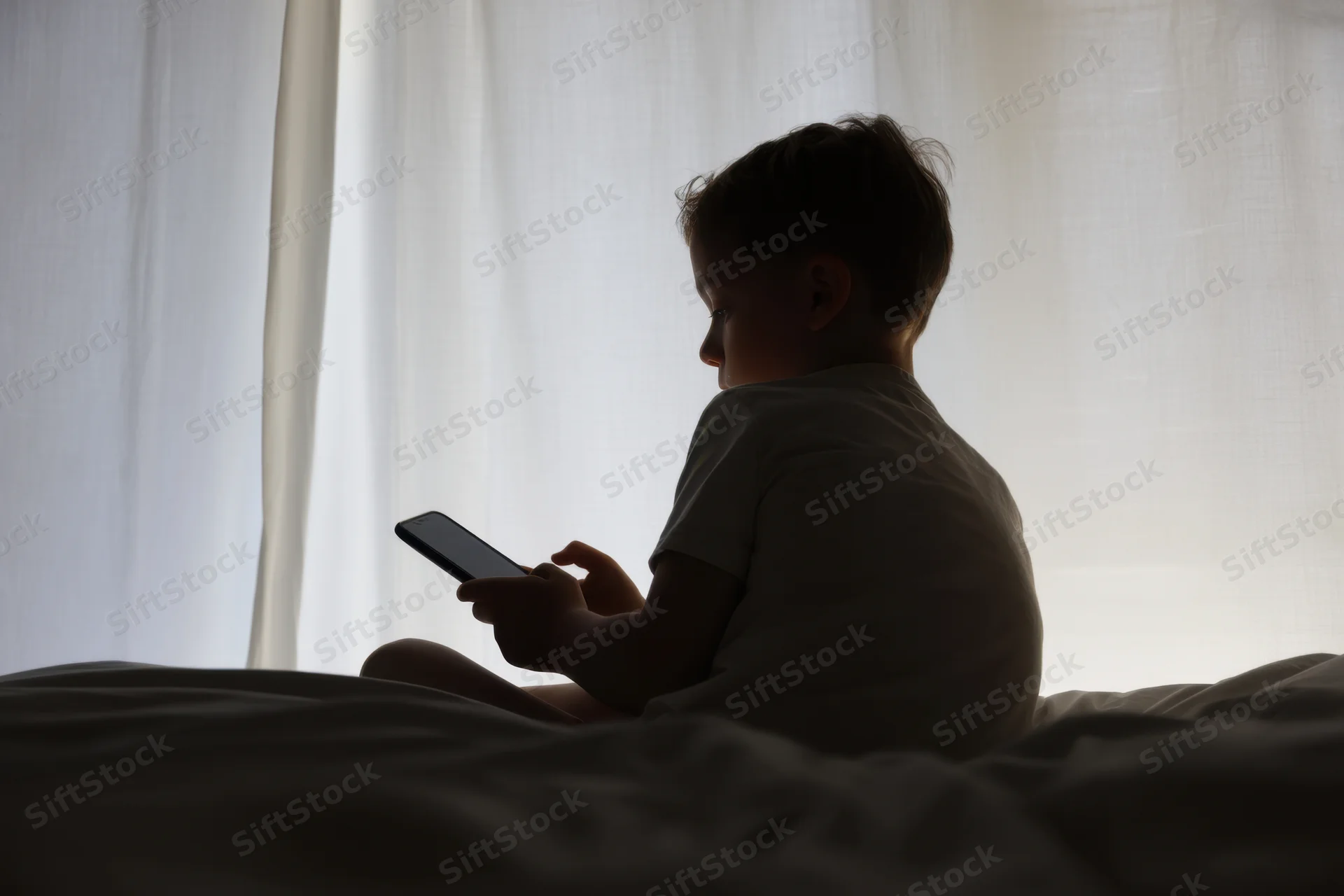Silhouetted child sitting on bed holding smartphone against window light
