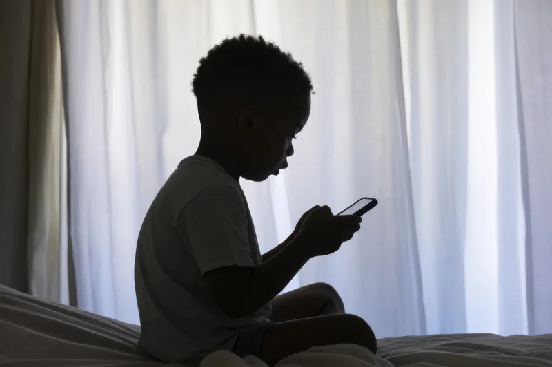 Silhouetted child sitting on bed holding smartphone against window curtain