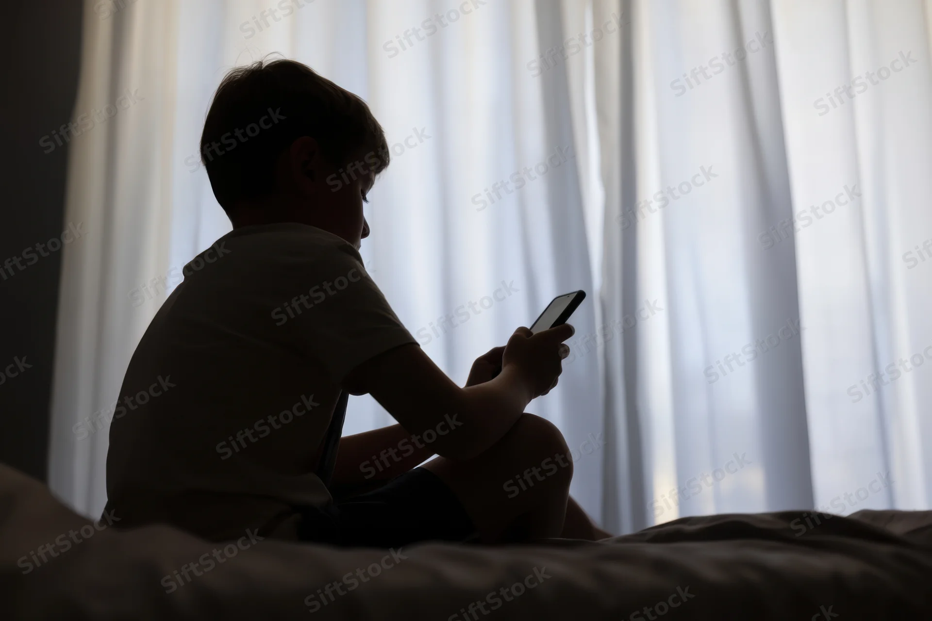 Silhouetted child sitting on bed holding smartphone in dimly lit bedroom near window