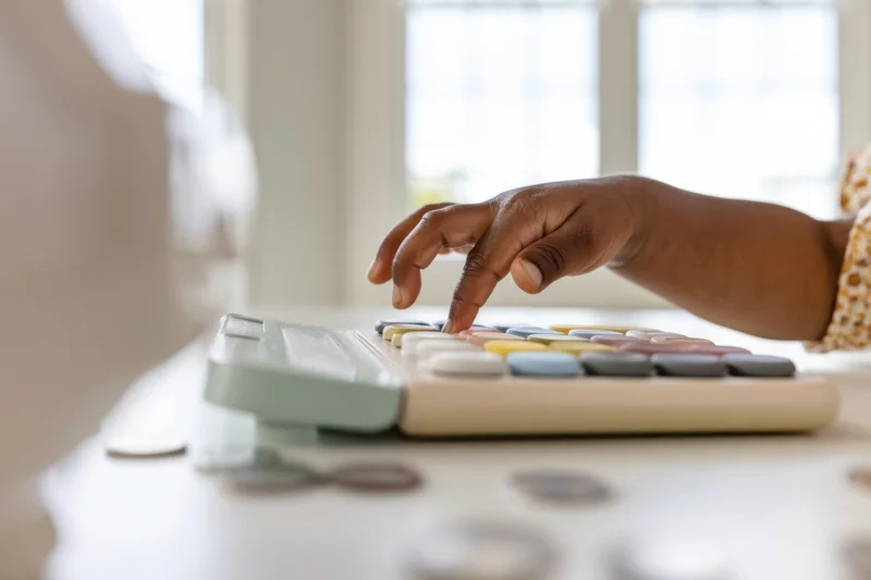 Child's hand pressing buttons on a pastel-colored calculator at a desk with coins nearby